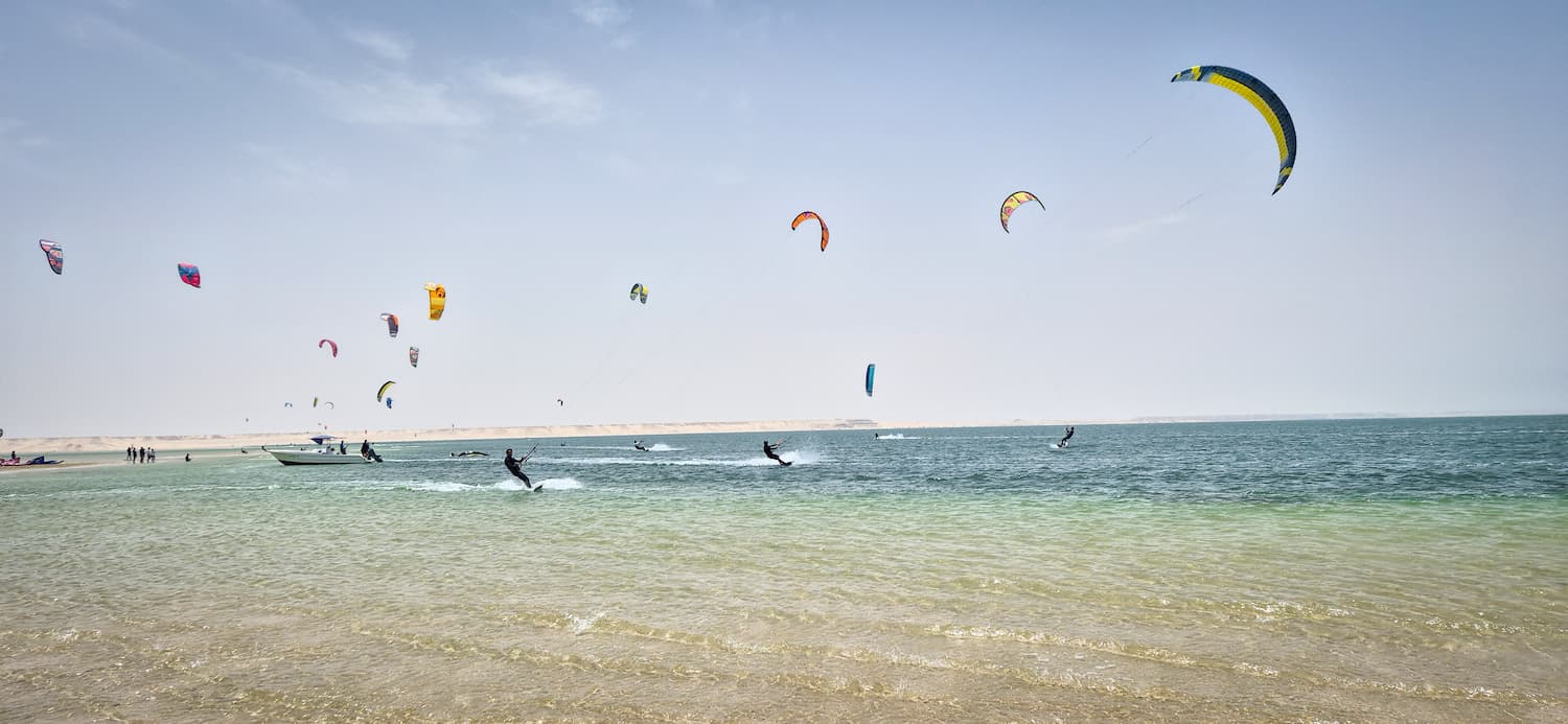 Masser af farverige kites på lagunen foran Auberge du Nomades du Sahara under en blå himmel