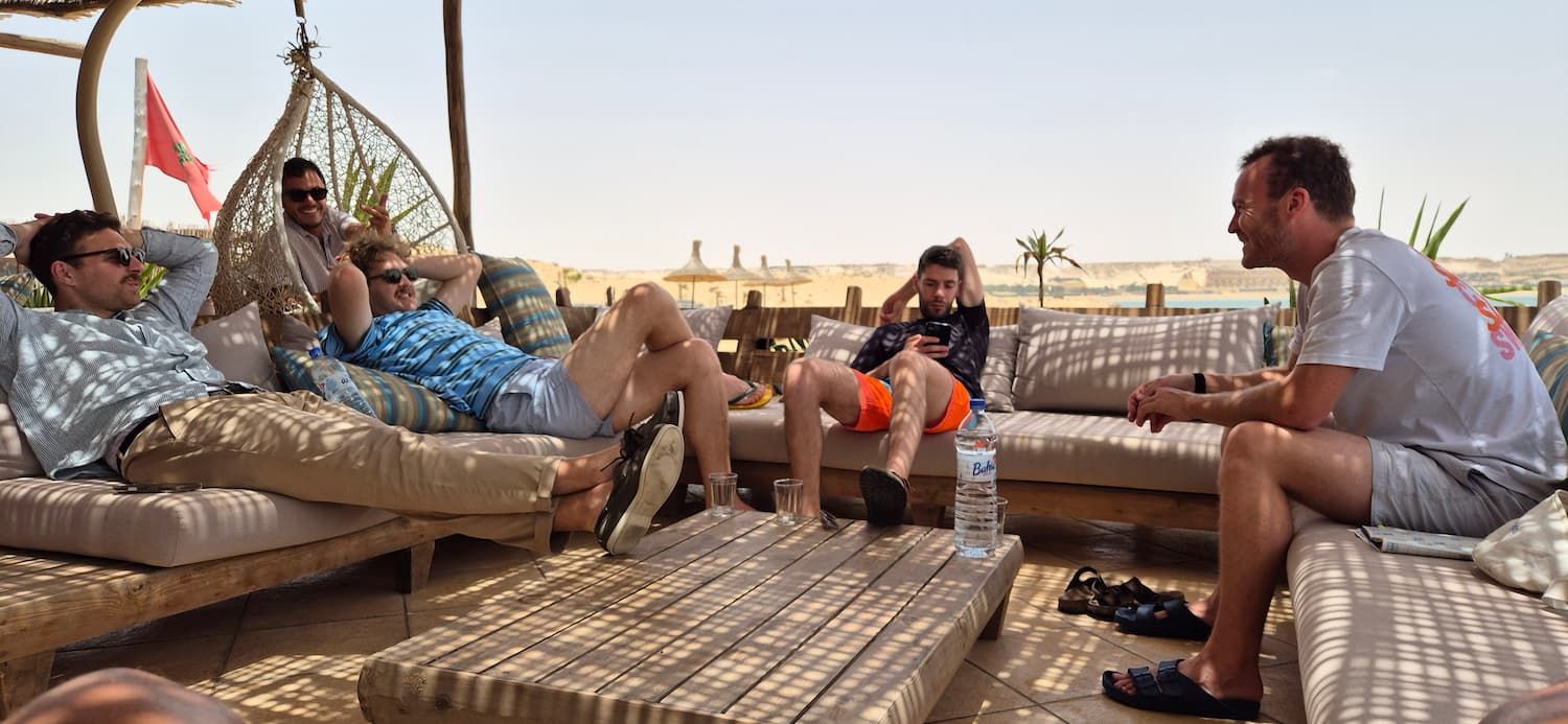 En gruppe glade kitesurfere der sidder henslængt og griner sammen i de store, hyggelige udesofaer på terrassen hos Auberge des Nomades du Sahara