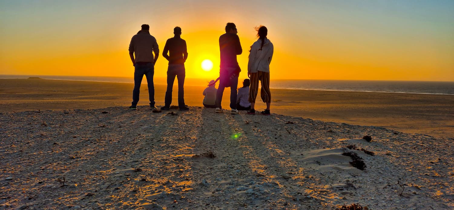 En gruppe kitesurfere står i silhuet på toppen af en sandbakke og ser en smuk, intens gul solnedgang over Dakhlas ørkenlandskab