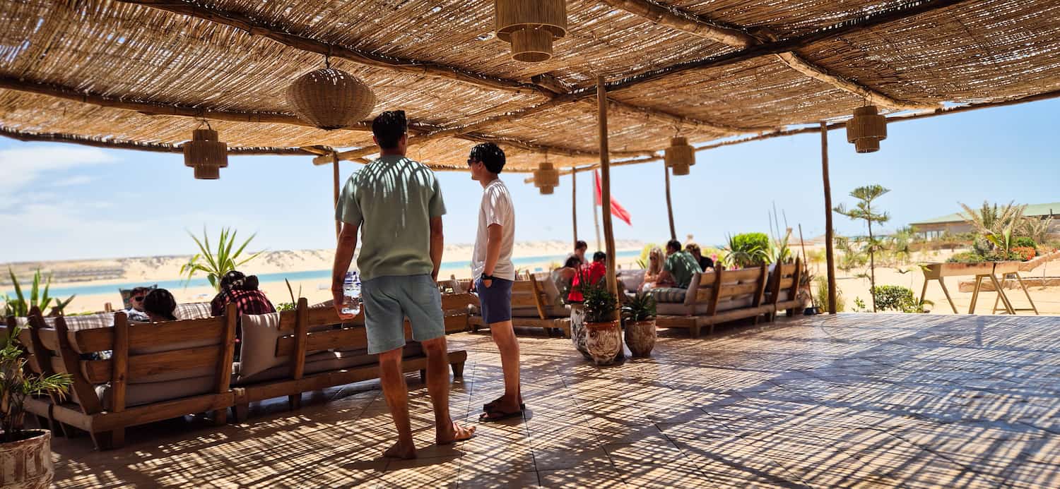 To mænd der snakker sammen på den skyggefulde stråtagsterrasse på Auberge des Nomades du Sahara med andre gæster siddende i baggrunden